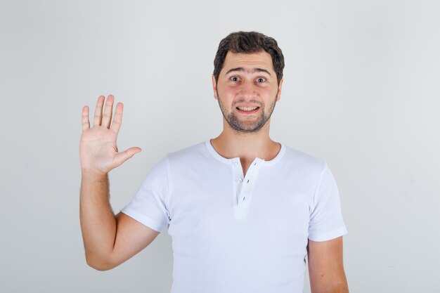 Young male waving hand in hello gesture in white t-shirt and looking jolly