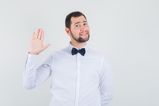 Young male waving hand to say hello or goodbye in white shirt and looking cheerful. front view.