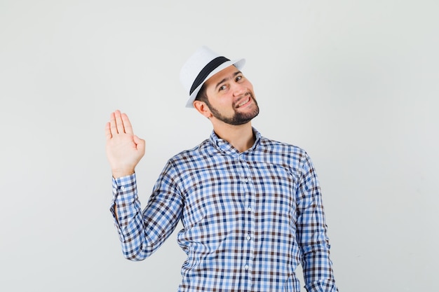 無料写真 チェックのシャツ、帽子、陽気に見えるで挨拶またはさようならを言うために手を振っている若い男性。正面図。