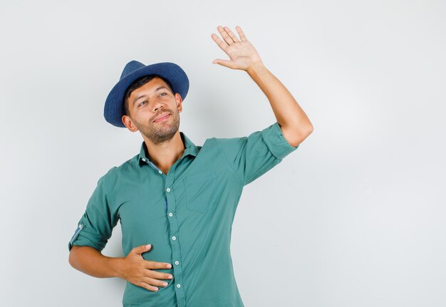 Young male waving at somebody in shirt, hat and looking glad.