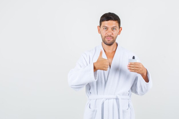 Young male in white bathrobe holding perfume with thumb up and looking positive , front view.