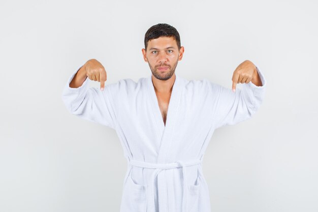 Young male in white bathrobe pointing down and looking serious , front view.