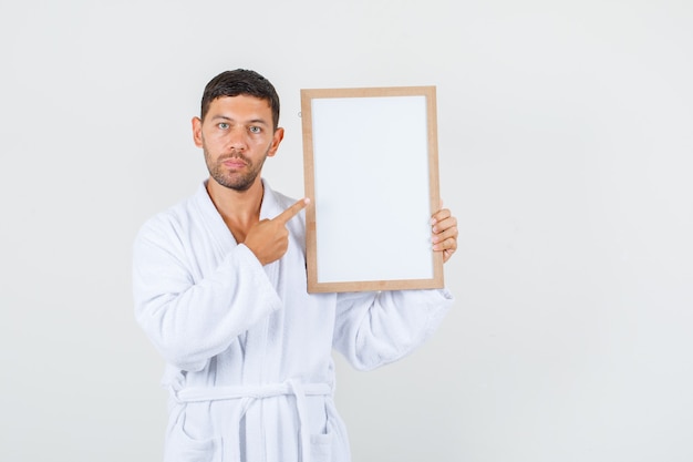 Young male in white bathrobe pointing at white board and looking strict , front view.
