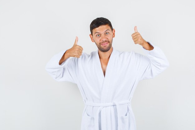 Young male in white bathrobe showing thumbs up and looking pleased , front view.