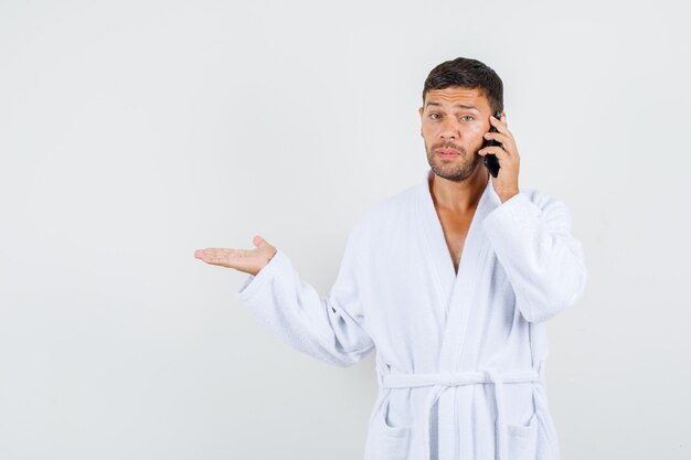 Young male in white bathrobe talking on phone with helpless gesture aside , front view.