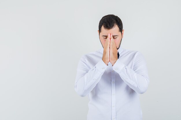 Young male in white shirt holding hands in praying gesture and looking hopeful , front view.