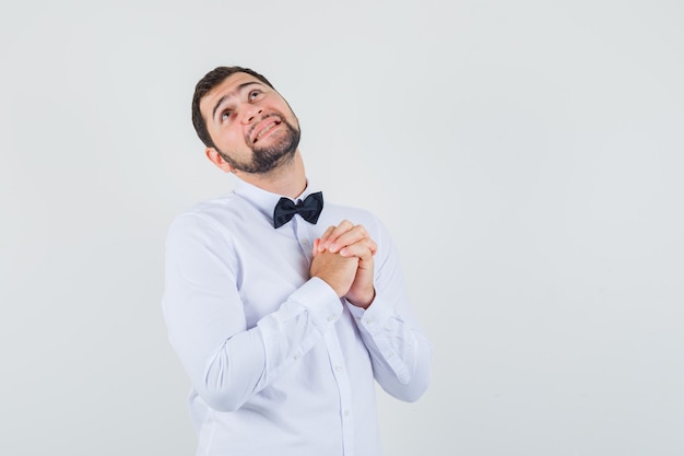 Young male in white shirt looking up with hands in praying gesture and looking dreamy , front view.