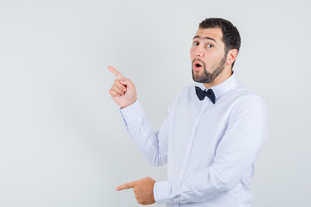 Young male in white shirt pointing fingers up and down and looking curious , front view.