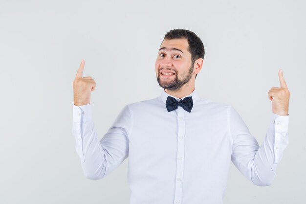 Young male in white shirt pointing fingers up and looking joyful , front view.