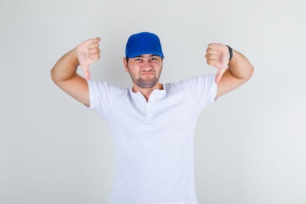 Foto gratuita giovane maschio in maglietta bianca, berretto blu che mostra i pollici verso il basso e sembra scontento