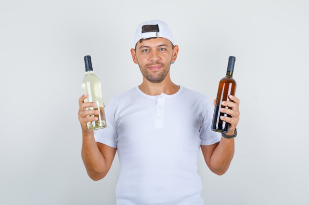 Young male in white t-shirt, cap holding bottles of alcoholic beverages and looking happy, front view.