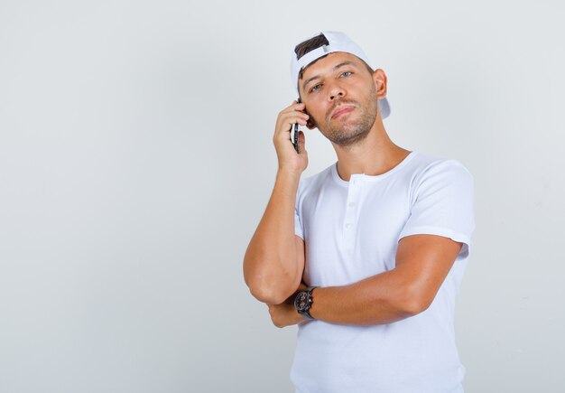 Young male in white t-shirt, cap holding phone and looking at camera, front view.