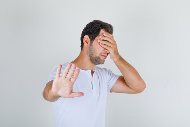 Young male in white t-shirt doing stop gesture and covering eyes