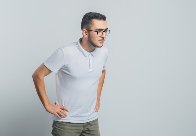 Молодой мужчина в белой футболке, штаны смотрит в сторону, положив руки на талию и выглядит сосредоточенным