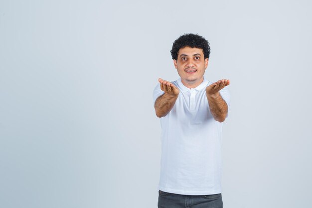 Young male in white t-shirt, pants making giving gesture and looking cheery , front view.