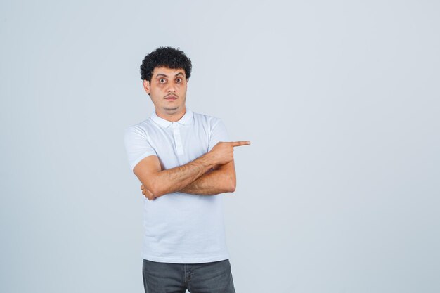 Young male in white t-shirt, pants pointing aside and looking surprised , front view.