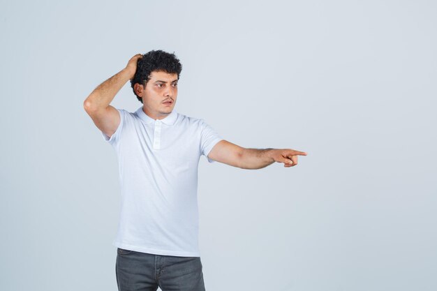 Young male in white t-shirt, pants pointing something away and looking puzzled , front view.