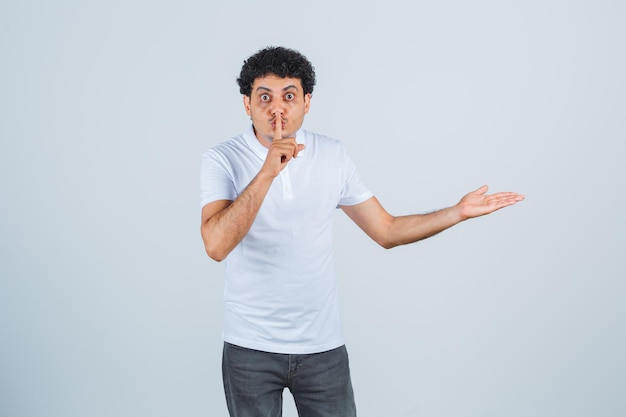 Young male in white t-shirt, pants showing something and silence gesture and looking careful , front view.