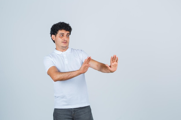 Молодой мужчина в белой футболке, штаны показывает жест стоп и выглядит испуганным, вид спереди.