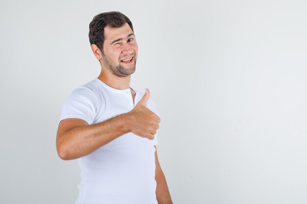 Young male in white t-shirt showing thumb up and blinking eye