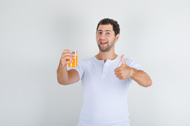 Young male in white t-shirt showing thumb up with glass of juice and looking happy