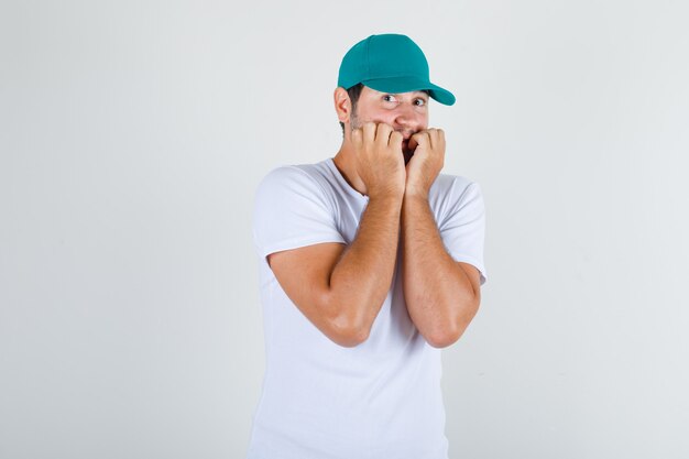 Young male in white t-shirt with cap keeping hands near mouth and looking scared