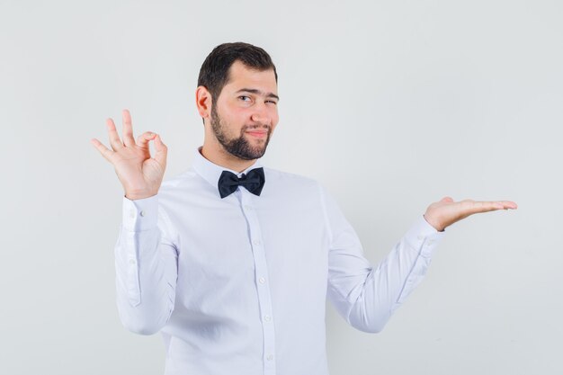 Young male winking eye, showing ok gesture, spreading palm aside in white shirt front view.