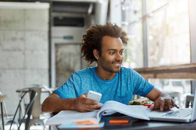 Free photo young male with dark skin and curly hair surrounded with books holding telephone in his hand looking in laptop with smile being happy to find what he needs for project. people, youth, education