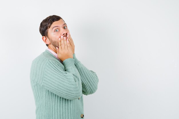 Giovane maschio con le mani sulla bocca in camicia, cardigan e dall'aspetto perplesso. vista frontale.