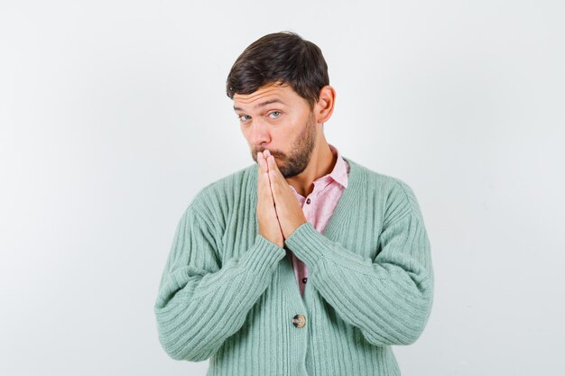 Young male with hands in praying gesture in shirt, cardigan and looking hopeful. front view.