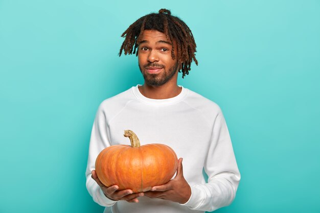 Young male youngster with dreadlocks, little beard, wears casual white sweater, holds orange pumpkin, prepares for holiday celebration