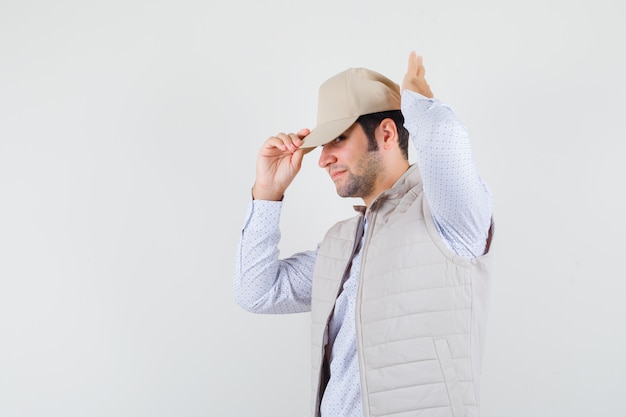 Young man adjusting cap while looking aside in shirt,sleeveless jacket and looking attentive .