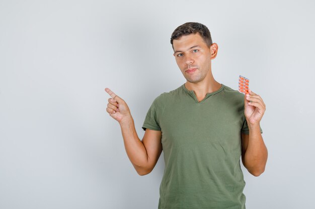 Молодой человек в зеленой футболке армии держит таблетки и указывая в сторону, вид спереди.