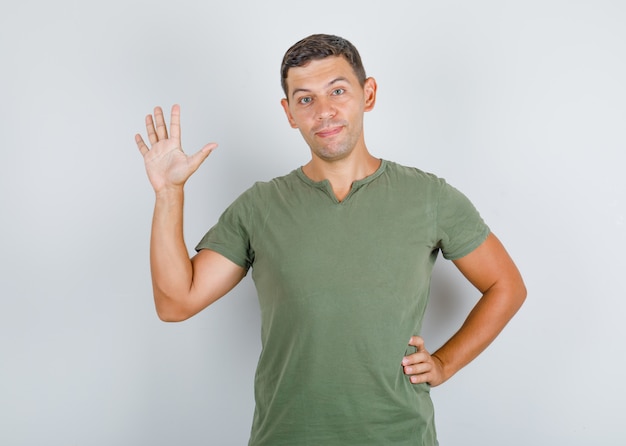 Young man in army green t-shirt raising palm in hello gesture and looking positive .