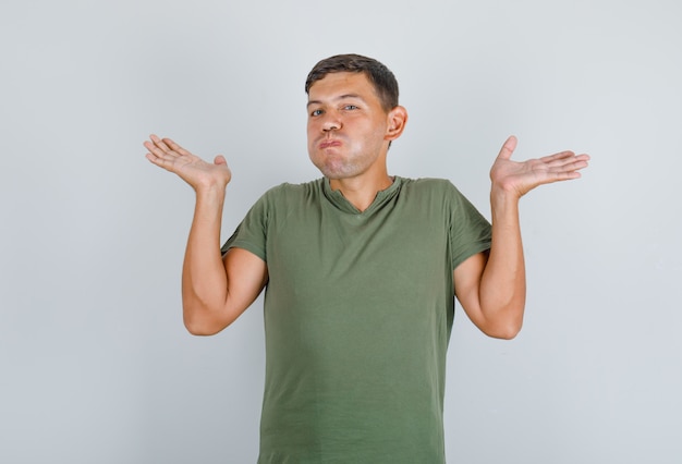 Young man in army green t-shirt showing helpless gesture with blowing cheeks, front view.