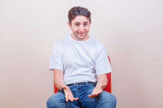 Young man asking something while sitting on chair in t-shirt, jeans and looking cheery