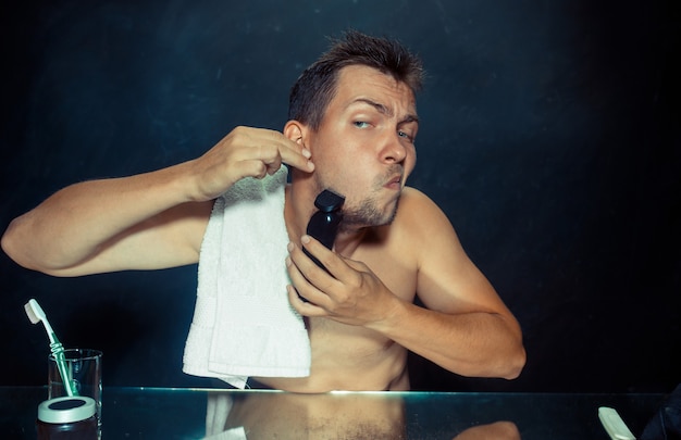 The young man in bedroom sitting in front of the mirror scratching his beard at home. Human emotions concept