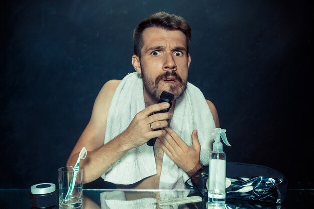 The young man in bedroom sitting in front of the mirror scratching his beard at home. Human emotions and lifestyle concept