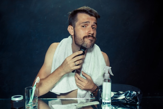 Молодой человек в спальне сидит перед зеркалом, почесывая бороду дома. Человеческие эмоции и концепция образа жизни