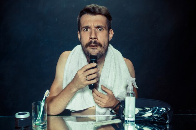 young man in bedroom sitting in front of mirror scratching his beard at home