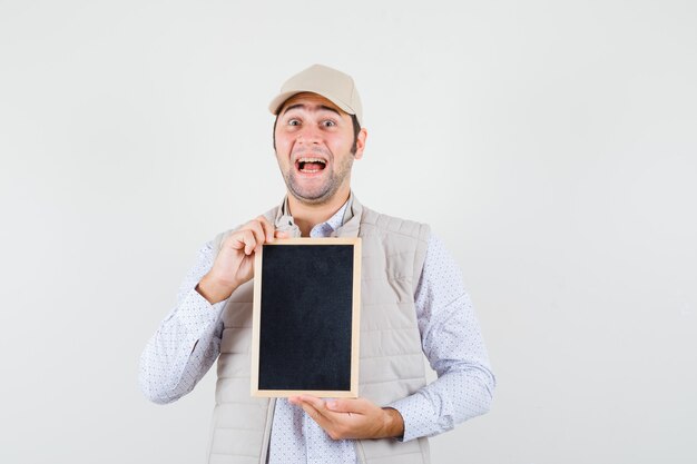 Young man in beige jacket and cap holding chalkboard and looking happy , front view.