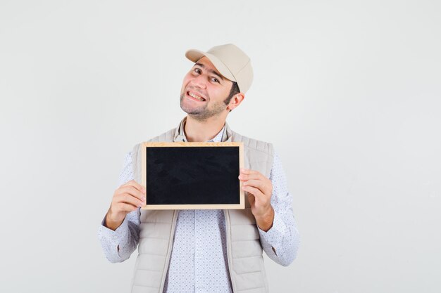 Young man in beige jacket and cap holding chalkboard and looking happy , front view.