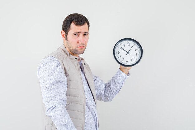 Young man in beige jacket and cap holding clock in one hand and looking dismal , front view.