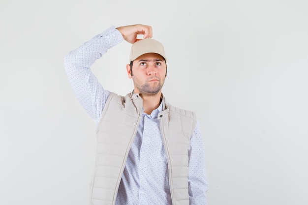 Free photo young man in beige jacket and cap holding hand on head, thinking about something and looking pensive , front view.