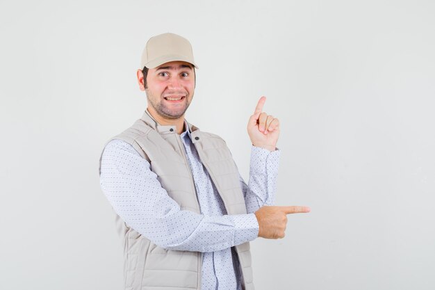 Young man in beige jacket and cap pointing right with index fingers and looking happy , front view.