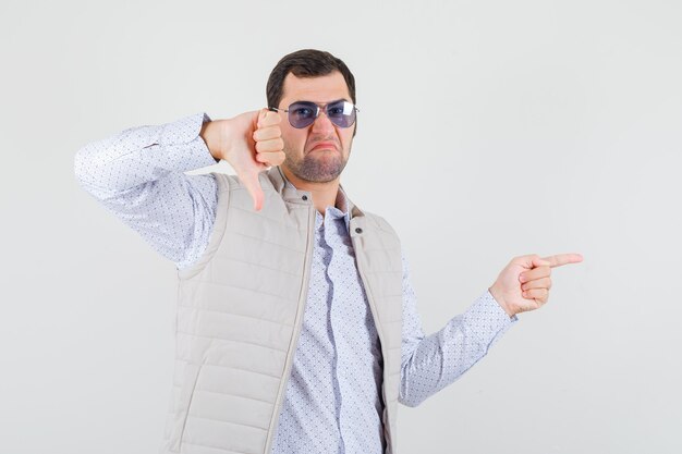 Молодой человек в бежевой куртке и кепке, надевая очки, указывая вправо указательным пальцем и показывая большой палец вниз и недовольный, вид спереди.