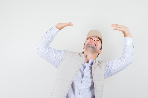 Young man in beige jacket and cap raising hands as holding something and looking exhausted , front view.