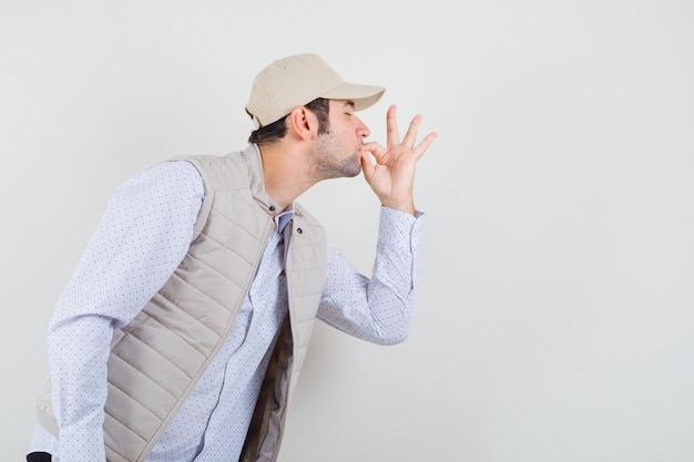 Young man in beige jacket and cap showing delicious gesture and looking happy , front view.