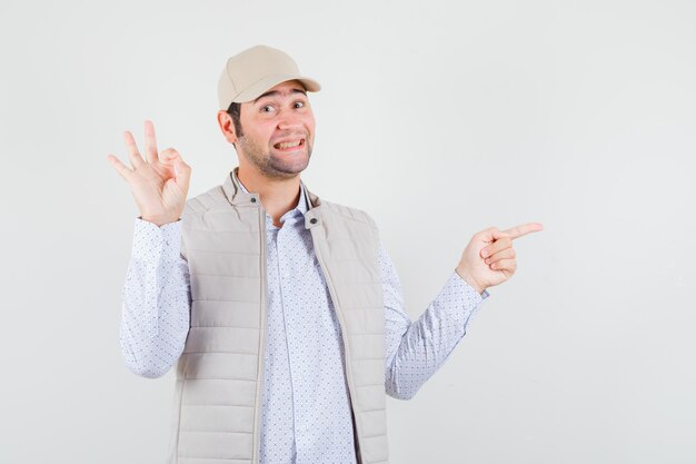 Молодой человек в бежевой куртке и кепке показывает знак ОК и указывает вправо указательным пальцем и выглядит счастливым, вид спереди.