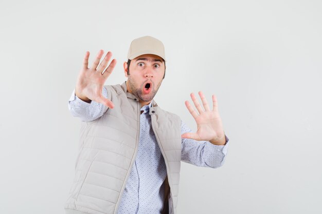 Young man in beige jacket and cap showing stop signs with both hands and looking surprised , front view.